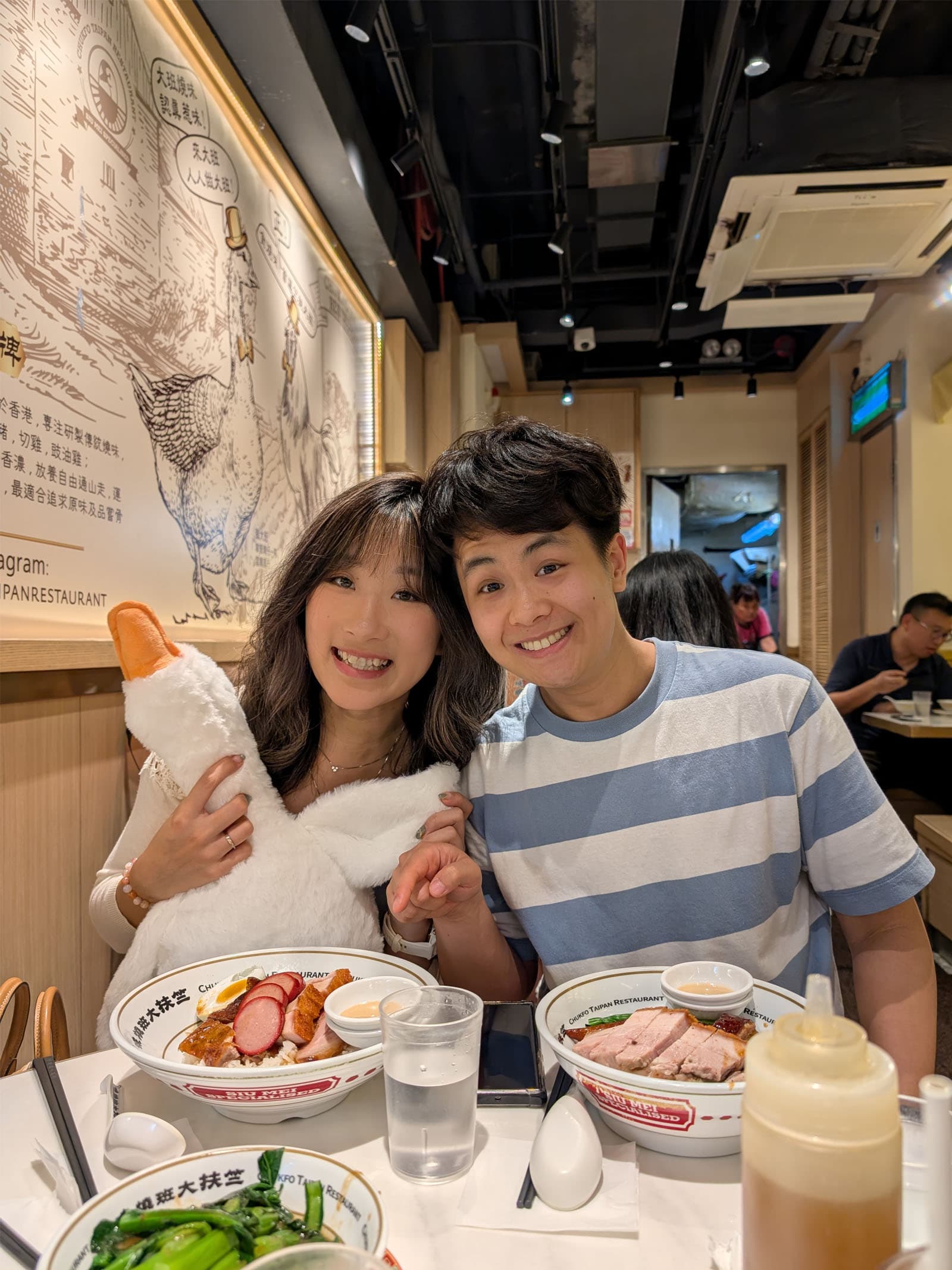 Thomas and Lylian holding a goose stuffed toy while wincing and pointing at two bowls of Hong Kong styled roasted goose BBQ rice in front of them.