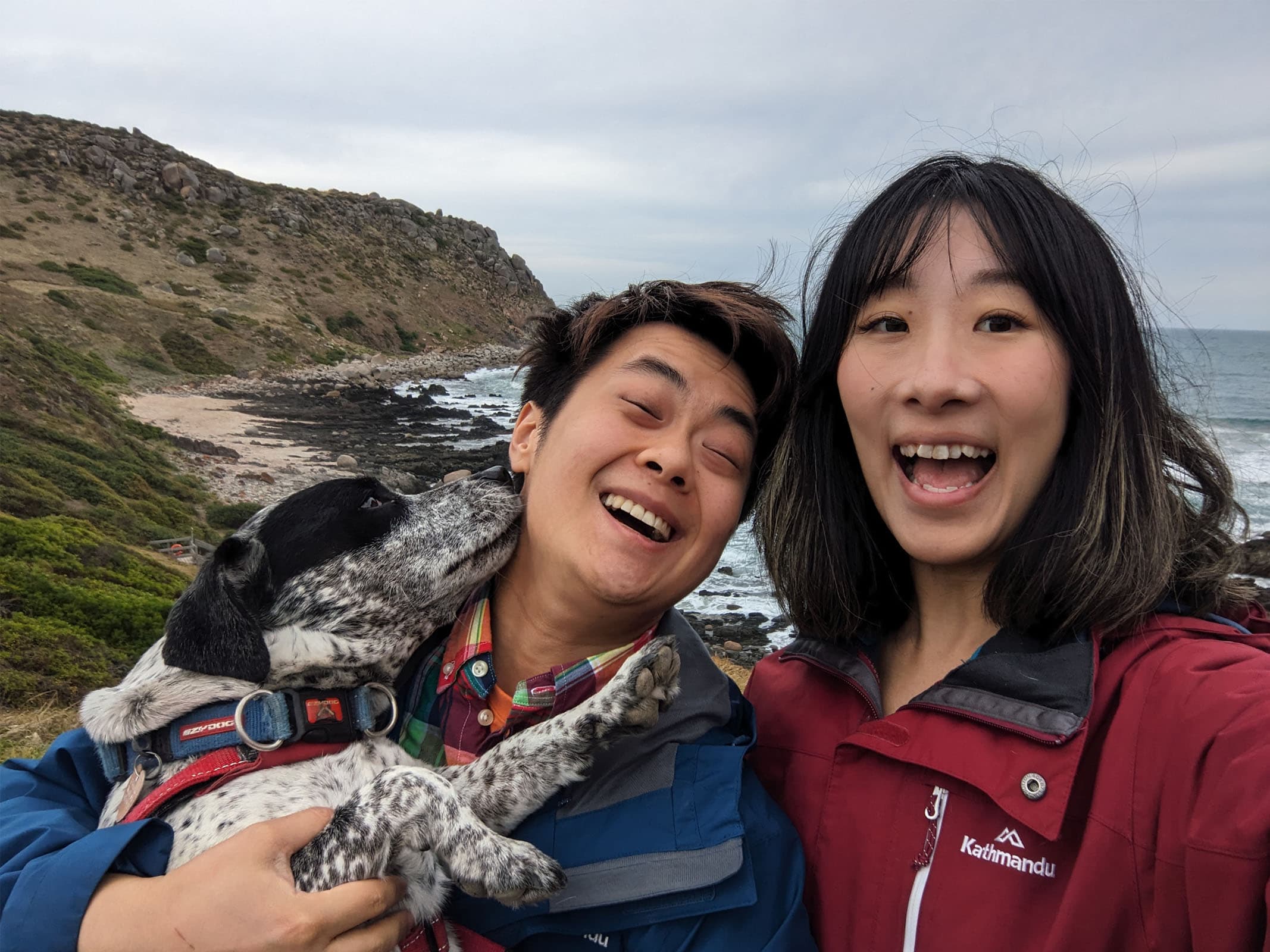 Thomas, Lylian and Moose taking a selfie at the coast of Victor Harbour on a cloudy day. Thomas is moving his head away while being licked by Moose.