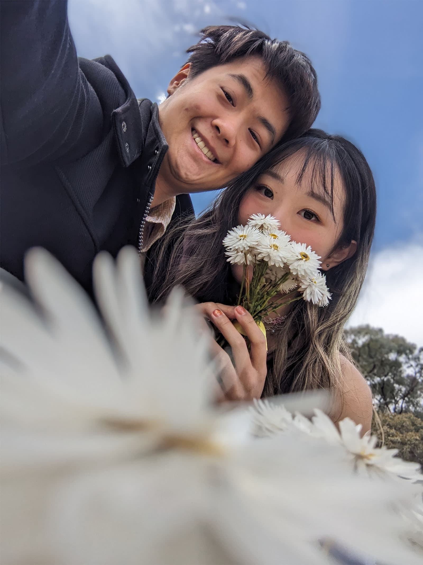Thomas and Lylian taking a selfie in a daisy farm. Lylian is hiding her face behind a bunch of flowers.