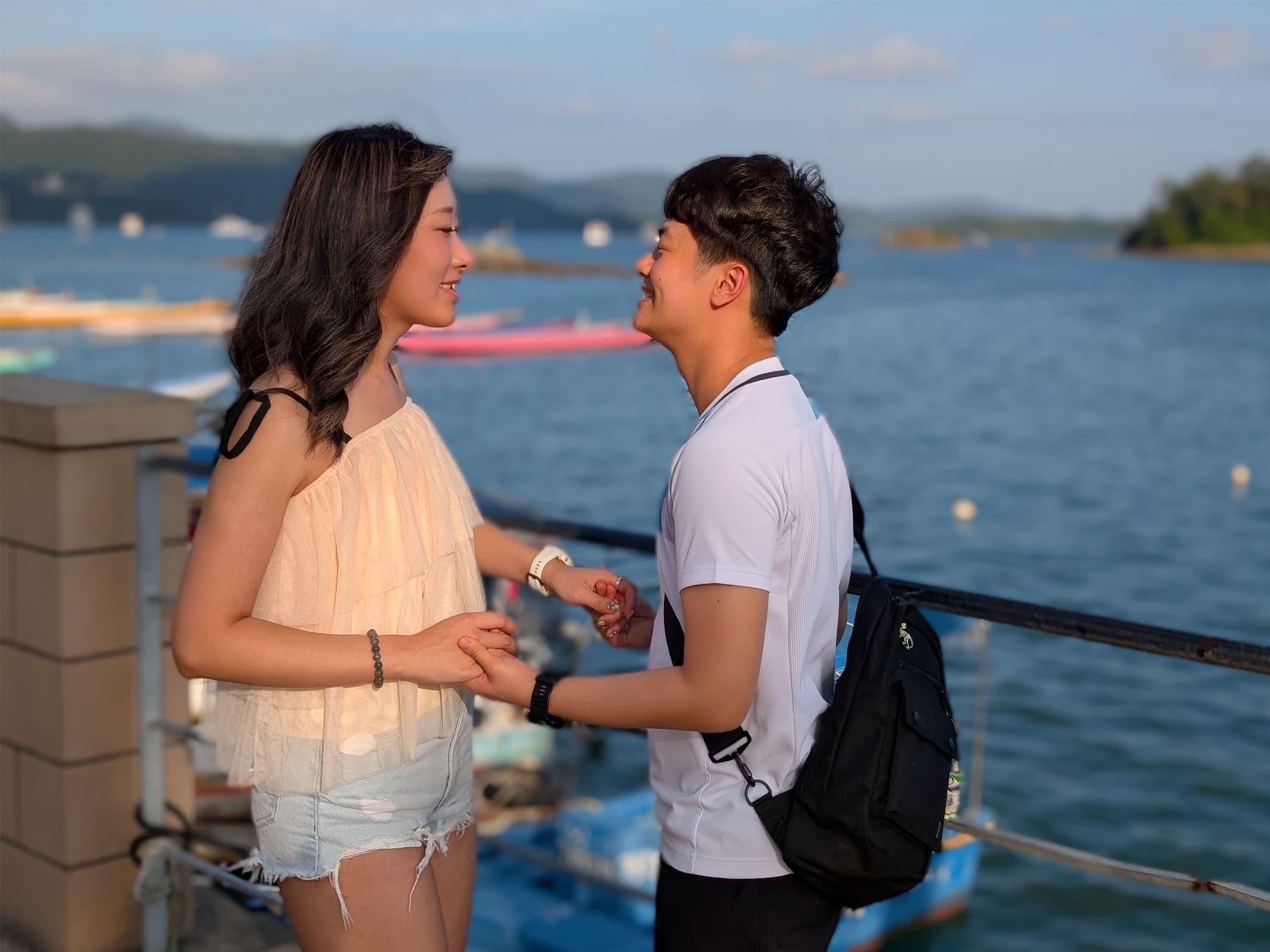 Thomas and Lylian holding hands and looking at each other at a small harbour in Hong Kong during sunset.