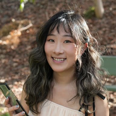Lylian smiling happily at the camera while holder her phone and wearing a peach-coloured summer top.